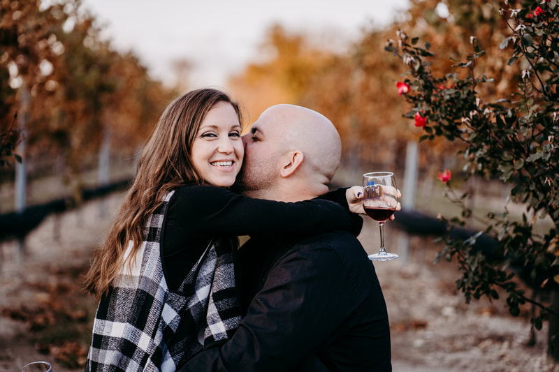 Fiance kissing bride to be on cheek in vineyard during the fall